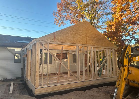 A new home addition framed and sheathed, with an excavator beside it and autumn trees behind