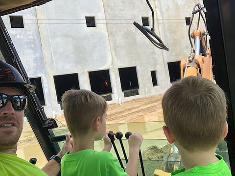 Allen in the cab of heavy equipment with two children, a concrete building behind them